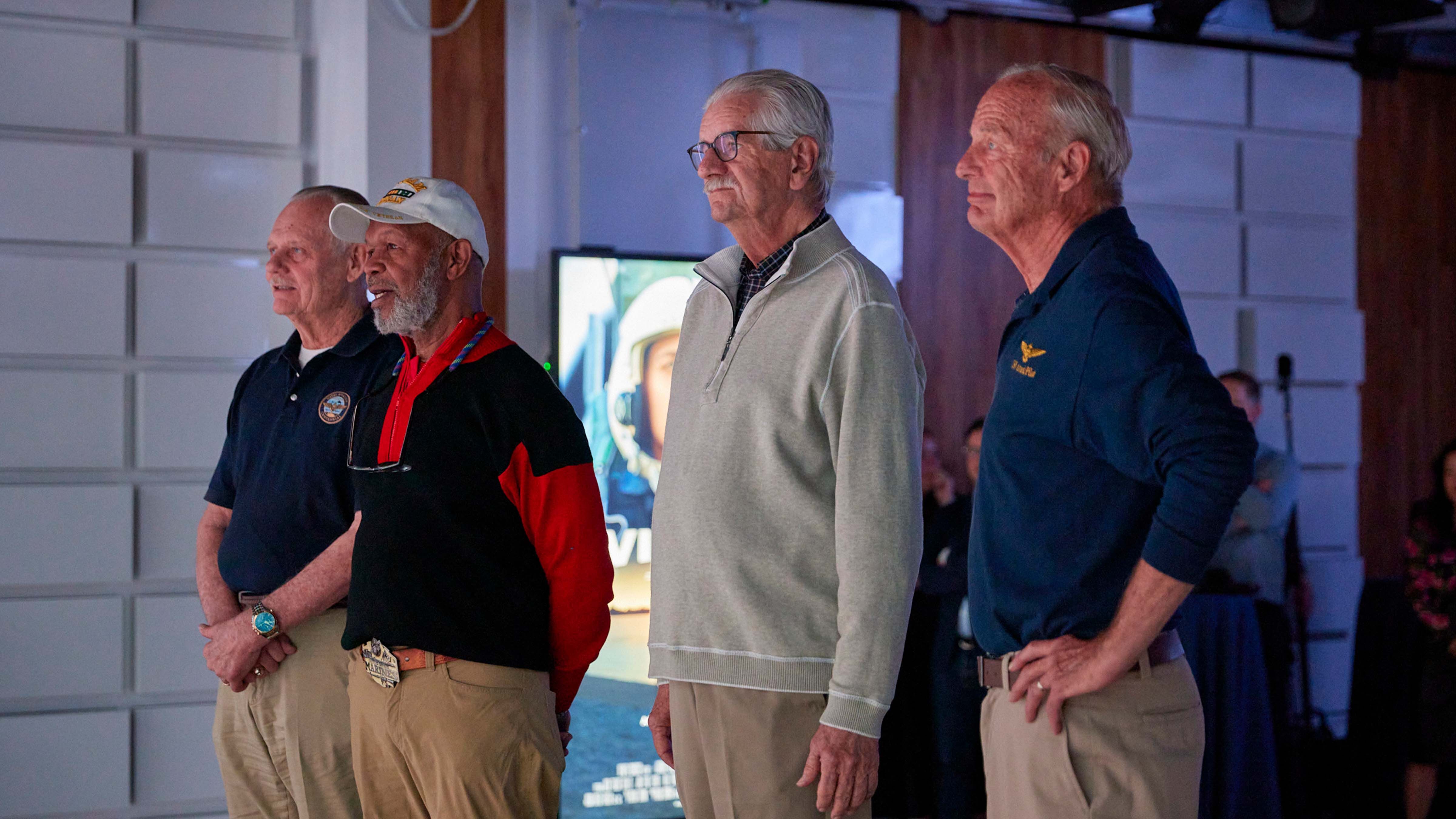 Four male army veterans, Jack 'Razor' Gillett, Elester 'Hollywood' Latham, Bill 'Offender' Moir, James 'Zeke' Lloyd watching a large movie screen at a film premier.